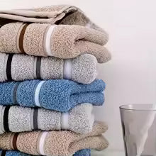 Stack of folded towels in beige, grey, and blue tones placed next to a glass on a white surface.